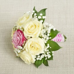 The bouquet before you gleams with the promise of a new beginning-a compact, tender arrangement that might grace a wedding morning in Brent Cross. Lush, cream-hued roses with velvety petals form the main cluster, their tranquil beauty reminiscent of quiet garden walks in the community's tranquil spaces. Breaking the gentle uniformity, a vibrant magenta-pink rose sparks joy in the heart of the bouquet, its colour as vivid as a sunset over local historic avenues. Wisps of delicate baby's breath flutter among the petals, creating an airy, cloud-like ambiance. Supporting the ensemble, broad, dark green leaves offer a grounding frame, lending both structure and contrast to the soft hues. The stem is wrapped in radiant pink satin ribbon, twined with a whisper of white pearls-a stylish touch inspired by the tastes of modern Brent Cross brides. Resting atop a lightly woven neutral fabric, the bouquet gives an impression of calm celebration and thoughtful artistry, ideal for a bridesmaid's hand or as a centrepiece at an intimate local gathering.