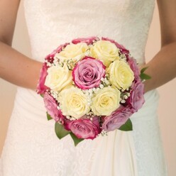 A masterpiece of local floral artistry, this Brent Cross bridal bouquet captivates with its thoughtful composition and radiant colours. Lush, creamy lemon-yellow roses nestle closely next to velvety fuchsia and pink roses, some petals fading into cool mauve or lilac, creating a layered interplay of hue and texture. Clouds of white gypsophila dance amidst the roses, their tiny blossoms softening the arrangement and adding a light, flirtatious touch. A few polished green leaves frame the bouquet's edge, enhancing its vibrancy and depth. The bride, her forearms bare and relaxed, holds the bouquet naturally just above the flowing fabric of her white wedding dress. The structured lacework of her bodice hints at timeless bridal tradition, while the flowing skirt, gently illuminated, lends an effortless grace. Every detail-from the carefully paired blooms to the airy filler-conveys the expertise and attention to detail that Brent Cross Flowers is known for in the local community. The light, uplifting mood echoes the atmosphere on a wedding morning in Brent Cross or Golders Green, where every arrangement is crafted to mark moments of happiness and new beginnings.