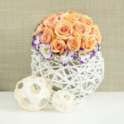 Set against a softly textured beige background, this refined centerpiece feels like it belongs on a beautifully laid table in Brent Cross, ready for a wedding breakfast or a special family celebration. A lush dome of peach and apricot roses crowns the arrangement, each flower fully open and luxuriant, their velvety petals layered tightly together so the cluster reads as a smooth, rounded form. The roses display gentle gradients of colour, from pale peach at the outer edges to warmer apricot tones deeper within, creating a sense of warmth and quiet joy. Circling the base of this rose globe is a halo of white lisianthus, each bloom edged with a striking violet or purple border that brings energy and contrast to the composition. The lisianthus petals are slightly ruffled, giving the arrangement an elegant, almost couture texture. Interspersed among these are small sprays of white gypsophila, their tiny blossoms like fine lace that softens and lightens the overall effect. The flowers are nestled into a bulbous, spherical vase woven from white rattan or wicker, its open latticework design letting glimpses of shadow and light play through the structure. This contemporary, handcrafted container calls to mind the stylish home décor pieces you might find in local Brent Cross boutiques or design-led stores in nearby Hampstead. Resting on the same bright white surface in front of the arrangement are two decorative spheres, one larger and one smaller, crafted from slender, pale wooden strips that mirror the woven texture of the vase. These orbs extend the natural, tactile theme and gently lead the gaze back toward the central bouquet. The whole scene communicates calm sophistication and romantic charm, perfect for depicting wedding flowers, modern home styling, or thoughtful floral gifts deliverable around Brent Cross.