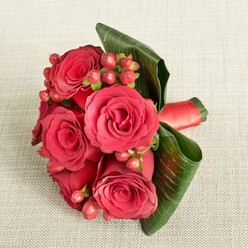 A compact, romantic bouquet of rich red roses lies gently on a pale beige, finely textured fabric, the kind of softly lit backdrop you might find in a calm sitting room off the high street in Brent Cross. The roses, around half a dozen in number, are fully open yet still plush, their velvety petals unfurling from tightly curled centres into layered whorls of deep crimson and scarlet that suggest warmth, devotion, and a touch of drama. Tucked between the blooms are clusters of small, rounded Hypericum berries in muted shades of red and reddish-pink, each berry like a tiny bead of lacquer, adding playful texture and brightness against the darker petals. Encircling the base, broad, dark green tropical leaves have been artfully folded and curved by an experienced florist, creating a natural, sculptural frame that supports the flowers and gives the arrangement a sleek, contemporary feel. A band of red satin ribbon grips the stems, its surface catching the light so that fine highlights run along the fold, hinting at the careful craftsmanship and attention to detail behind the design. The gentle, even lighting reveals every contour and curve-the veins in the leaves, the shadows between petals, the shine on the berries-without overwhelming the composition, allowing the rich colours to speak for themselves. This bouquet feels equally suited to being carried by a bridesmaid walking through a ceremony near Brent Cross Shopping Centre, placed as a romantic centrepiece on a dinner table, or delivered as a heartfelt gesture of love and gratitude to a home in the surrounding North London neighbourhoods.
