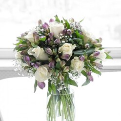 A sophisticated glass vase holds a carefully composed bouquet of white roses, lavender alstroemeria, and delicate baby's breath, illuminated by soft natural light that suggests it is resting near a bright window in a Brent Cross flat or townhouse. The roses are fully open yet still crisp at the edges, each petal curling back in smooth, creamy layers that give the arrangement a luxurious, almost satin-like texture. Nestled around them, the alstroemeria blooms shift gently from pale lilac to muted purple, with fine, darker markings running along their inner petals, as if painted by hand. Some of these Peruvian lily flowers are still in bud, enclosed by green sepals that hint at blossoms yet to unfurl, adding a sense of movement and quiet anticipation. Wisps of white baby's breath appear to float between the larger heads, forming a lace-like veil that softens the transitions from bloom to bloom and lends the design an ethereal, clouded quality. Lush greenery surrounds and supports the flowers: broad, dark leaves anchor the base visually, while slimmer stems and shoots reach upward, shaping the bouquet into a generous, rounded dome. The clear cylindrical vase reveals the straight, aligned stems below the waterline, their fresh cut ends and subtle colour variations showing the care of an experienced local florist familiar with the tastes of families along the North Circular and around Brent Cross station. The background is bright yet blurred, perhaps a light wall or sheer curtain, so that the focus remains solely on the interplay of white, green, and purple. This arrangement feels calm, respectful, and uplifting at once-a design that could just as easily stand on a polished dining table for a quiet celebration as on a bedside in nearby Cricklewood, bringing solace, admiration, or a gentle message of hope to whoever receives it.