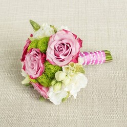Resting gracefully on a pale beige, woven fabric that resembles finely textured linen, this small hand-tied bouquet captures the feeling of a quiet, joyful moment before guests arrive at a Brent Cross celebration. Three pink roses form the heart of the design, their blossoms open and full yet still slightly cupped at the centre, revealing intricate curls of petal within. Each rose shifts gently from a soft pastel blush at its core to deeper, almost fuchsia tones on the outer edges, giving the flowers a painterly, layered depth. Between and around the roses, vibrant lime-green button chrysanthemums create rounded, pom-pom clusters that contrast beautifully with the smooth rose petals, adding a cheerful, modern pop of colour that feels at home in a contemporary North London apartment or a stylish venue off the A41. Creamy white and ivory filler blooms, most likely lisianthus or freesia, thread their way through the bouquet, their delicate, fluttering petals softening the overall palette and bringing a sense of purity and lightness. Tucked discreetly among them are tiny white star-like flowers with yellow centres, similar to waxflower, which introduce a fine, textured detail you might only notice when holding the bouquet in your hands. The stems are neatly bound together with a vivid fuchsia pink satin ribbon, smooth and lustrous, wrapped with care to form a comfortable handle. A fine strand of white faux pearls spirals across the ribbon, intersecting at gentle angles, adding a refined, bridal touch without overpowering the flowers. Under soft, even lighting, the colours glow naturally, suggesting a bouquet designed for a bridesmaid, prom, or engagement shoot around Brent Cross, created to be admired both up close and from across the room.