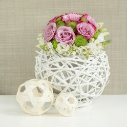 Captured in soft, flattering light, this floral centerpiece feels like it is already in place on a wedding table in Brent Cross, waiting for guests to sit down and clink glasses. The bouquet is shaped into a low, domed profile, perfect for conversation, with lush pink and lavender roses forming the main body. Each rose is generously open, their velvety petals layered in gentle spirals, some tipped with deeper pink tones that hint at a romantic, almost vintage charm. Between these, bright green button chrysanthemums nestle closely, their small, rounded heads adding a fresh, modern edge and enlivening the pastel palette with their vivid, grassy hue. At the base, tiny white filler flowers-delicate and cloudlike-peek out from beneath the larger blooms, creating a soft fringe that gently breaks the line between flowers and container. The arrangement sits within a white spherical vase woven from slim, rattan-style twigs, forming an airy, openwork structure that suggests handcrafted artistry while still feeling chic and contemporary. It resembles the kind of decor piece you might discover in a well-curated Brent Cross boutique or stylish home overlooking the North Circular. To the left on the same white surface, two pale wicker spheres lie side by side, one noticeably larger than the other, their lattice-like design mirroring the vase and lending the composition a sense of repetition and harmony. Behind, a softly focused beige background, similar to natural linen, provides gentle texture without distraction. The overall effect is serene and polished, with even lighting that illuminates each bloom, petal edge, and woven detail, making this centerpiece a beautiful choice for wedding receptions, anniversary dinners, or refined celebrations across Brent Cross and the surrounding area.