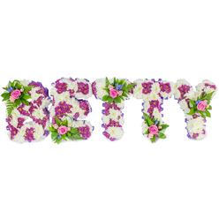 When someone dear is remembered with a name spelled in flowers, this tribute reads as both statement and whisper: the letters BETTY are formed in substantial, freestanding blocks whose main surfaces are a mass of soft white chrysanthemums, their cushiony texture catching soft light and offering a sense of peace. Within each letter a border and internal motif of vivid purple-magenta chrysanthemums create definition and a gentle visual heartbeat, like a melody threaded through a quiet hymn. Edges are carefully wrapped in subtle purple ribbon that gives the composition a composed, tactile outline, while small posies sit like quiet gestures-on the B's upper curve, atop the central T, and on the Y's branching arm-composed of blush-pink roses in mixed stages of bloom, tiny bright blue flowers that may be delphiniums or cornflowers, and sprigs of fern and fresh greenery. These clusters bring an intimate, garden-like softness to the otherwise structured lettering, the roses' faint fragrance imagining a comforting presence among mourners. Set against a pristine white backdrop, every hue and texture reads clearly: the creamy whites are tranquil, the purples are reverent, the pinks are tender, and the blues are slightly hopeful. A memorial piece like this is appropriate for services at local venues or crematoria near Brent Cross and resonates with families from nearby Golders Green and Dollis Hill who prefer a personalised floral tribute that balances dignity, craftsmanship, and heartfelt remembrance.
