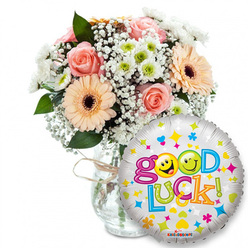 A carefully composed 'Good Luck' ensemble, styled with the sensibility of a local Brent Cross florist and set on a bright white background that lets every hue read clearly. On the left, a lush, hand-arranged bouquet rests in a clear glass vase finished with a rustic twine knot; powder-pink and soft-peach roses create a romantic, tender centre, their layered petals suggesting a gentle, powdery scent. Among them sit warm peach-orange Gerbera daisies with their bold, dark brown centres - petals smooth as silk and energetically open - while generous clouds of white gypsophila weave a lace-like softness through the stems. Small white and lime-green chrysanthemum clusters pop like tiny stars among the foliage, lending crisp contrast and a fresh, grassy note. Dark green leaves peek through, providing structure and depth. In the foreground to the right, a shiny silver foil balloon overlaps the bouquet, its playful Good Luck! message spelled in buoyant, multicoloured letters; the two o's have been replaced by sunny yellow smiley faces - one winking, one sticking out its tongue - and the balloon is showered with tiny heart and star motifs in pink, blue and yellow, with a discreet KALEIDOSCOPE mark at the base. The overall light is soft and even, the glass reflecting a subtle sheen that emphasizes freshness. As a floral offering from Brent Cross Flowers, this pairing feels personal and encouraging - a perfectly balanced gift for farewells, exam support, interviews or new beginnings across Brent Cross, Cricklewood and nearby Golders Green.