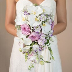 Visualize a bride set against a tranquil, softly blurred beige backdrop, her arms elegantly cradling a luxurious bridal bouquet lovingly composed by a seasoned florist in Brent Cross. The bouquet demands attention with its harmonious blend of pure white, soft pink, and gentle lavender flowers cascading naturally downward in a timeless teardrop shape. Central to the arrangement, large white phalaenopsis orchids with golden centres unfurl their velvety petals, radiating a refined grace. Around them, garden roses in the faint blush of a morning sky at Brent Reservoir evoke classic romance, while clusters of pale lavender stock flowers lend subtle spikiness reminiscent of wild blossoms bordering local woodlands. Scattered between, white lisianthus ruffles edged in a violet-pink hue add artistry, and weightless clouds of baby's breath enhance lightness and breadth. Flecks of greenery peek through, tying all elements together with a gentle touch. The bride's gown, strapless and textured with antique ivory lace, supports the bouquet's abundant beauty, focusing every eye on this expressive symbol of love and happiness. The entire composition tells a sensory-rich story-fragrant, textural, and full of heartfelt anticipation, beautifully reflecting the refinement and community character of Brent Cross weddings.