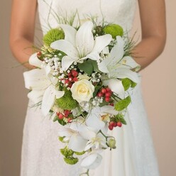 Bathed in radiant daylight, a bride's hands gently cradle a lavish, teardrop-shaped bouquet-a floral masterpiece reflecting the heartfelt hospitality and style of Brent Cross. The focal point is a starry cluster of white lilies, each petal traced with warm freckles, bringing together bold structure and a feather-soft feel. A creamy rose, its petals tight and romantic, glows at the center, exuding classic beauty. Around these central blooms dance moss green pom-pom mums, whose vibrant color and plush texture evoke the verdant spirit of Brent Cross's lush open spaces. Tiny white clouds of baby's breath break up the brightness, imbuing the bouquet with a weightless, ethereal quality. Striking red hypericum berries, with their berry-bright luster, are artfully interspersed, hinting at celebration and local community warmth. Cascading downward, elegant white phalaenopsis orchids trail with effortless grace, suggesting the gentle curves of the area's leafy avenues. Fine green foliage, both structured and wispy, frames and elevates the arrangement, while delicate strands seem to sway with each movement. The background is softly blurred, letting every detail shine against the lace and satin of the bride's gown-a scene that could unfold in an elegant Brent Cross venue or an intimate gathering at a nearby home. This bouquet tells a story of hope, artistry, and the joyful promise of a new beginning, inspired by the vibrant floral heritage of North West London.