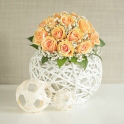 A refined floral display captures the eye: a domed bouquet of approximately twenty to thirty soft peach roses, each one layered with tightly swirled petals that deepen to a rich apricot at the center and fade to a gentle cream at the edges. The roses are pressed closely together, forming a rounded cushion of colour that feels warm and embracing, like the late afternoon light spilling over Brent Cross Station. Between the roses, clusters of tiny white gypsophila blossoms shimmer like a halo of fine mist, their airy structure subtly softening the overall shape. Occasional dark green leaves nestle at the base, adding contrast and reminding you that these are fresh, living stems. The arrangement is cradled in a striking white spherical vase, created from strands of woven string or wicker that twist and cross to form an open, airy cage. This delicate lattice orb rests solidly on a sleek white surface, perhaps a dining table in a newly styled flat off the A41, ready to welcome guests. In front of the vase, two smaller decorative orbs echo the same crafted texture: the larger one patterned like a minimalist football, the smaller more freely tangled, both in harmony with the main container. A pale beige or greige wall provides a calm, uncluttered backdrop so the roses truly glow. The soft, even lighting gives the impression of natural daylight filtering through a nearby window, making the scene feel inviting, intimate, and expertly composed, ideal for a wedding reception, anniversary dinner, or stylish home staging in and around Brent Cross.