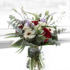 Bathe in the gentle, reflective light reminiscent of mornings along Brent Cross's leafy avenues, this bouquet exudes serene elegance and comforting warmth. Deep burgundy roses, velvety and lush, provide unmistakable drama, their allure enhanced by the pure white of gerbera daisies, each flower poised with a dark, expressive center. Twining among them are mauve and lavender lisianthus, half-opened buds hinting at new beginnings-perfect for marking personal milestones or expressing heartfelt sentiments to friends in Golders Green. Round leaves of eucalyptus offer a silvery-green coolness, framing the more vibrant blossoms and infusing the display with a soft, invigorating aroma. Clouds of white filler flowers nestle alongside broad, structured leaves that cradle the bouquet in sculptural fashion. Glittering green stems reveal the care behind every selection, submerged in the glass vase, with sunlight dancing across the arrangement. The subtle interplay of rose and eucalyptus fragrance transports the spirit to local gardens near Brent Reservoir, making this bouquet an inspired gift for any occasion, crafted by the expert hands at Brent Cross Flowers.