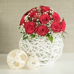 This image presents a romantic floral composition that feels perfectly at home on a wedding table in Brent Cross, just a short walk from the shopping centre's bustling streets. At its centre, a dome-shaped bouquet of deep red roses rises from the table, each bloom fully unfurled with plush, velvety petals that curve inwards to form a hypnotic spiral. The roses are massed tightly together, creating a saturated field of crimson that speaks of love and celebration. Between these passionate tones, clouds of tiny white baby's breath weave gracefully, their delicate, star-like flowers softening the intensity and lending the arrangement a dreamy, almost ethereal quality. Small highlights of green foliage peek from within, adding a hint of freshness and grounding the bouquet in nature. The flowers rest in a distinctive white vase, round and bulbous, crafted from a network of interlaced strands that cross and twist into an open, basket-like lattice. The structure feels both modern and artisanal, much like the curated decor found in contemporary Brent Cross apartments. Set upon a smooth, white tabletop are two decorative woven spheres in creamy off-white tones, one larger, one smaller, their loose open weave mirroring the pattern of the vase and adding gentle visual rhythm. Behind the composition, a softly textured wall in pale beige fades quietly into the background, ensuring there is nothing to distract from the rich colours and intricate forms of the flowers and decor. The overall impression is of polished yet intimate elegance, a centrepiece well-suited to wedding receptions, engagement dinners, or romantic gatherings across Brent Cross and the surrounding North London neighbourhoods.