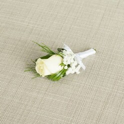 A classic white rose boutonniere is presented on a gentle angle, resting against a pale beige, linen-like cloth that recalls the soft neutrals of a well-dressed Brent Cross wedding venue. At its heart, a single rosebud in creamy white, shaded very slightly toward ivory at the center, is captured just as its petals begin to open, offering the impression of quiet promise and new beginnings. The bloom is plump yet refined, with smooth, velvety petals that catch the soft, natural light and show faint shadow along each fold. Surrounding this focal flower, delicate clusters of white baby's breath form a halo of tiny, rounded blossoms, their airy presence softening the arrangement and adding a cloud-like texture, much like the light mist that can hang over the paths near Brent Reservoir on an early morning. Slender, rich green leaves and a few fern-like sprigs frame the rose and gypsophila, offering contrast and structure, their curves and points guiding the eye along the length of the boutonniere. The stems are expertly bound with a pure white satin ribbon, its surface smooth and gently lustrous, finishing in a small, precise bow that sits just under the bloom, hinting at the skilled hands that tied it. The neutral background features a visible woven pattern, creating a calm, organic setting that allows the white florals and greenery to stand out without distraction. Soft, diffused lighting from above picks up the subtle shadows in the petals, the crisp edges of the leaves, and the gentle sheen of the ribbon, evoking a sense of quiet, polished elegance. This image would suit content on groom's attire, traditional lapel flowers, or refined wedding details for ceremonies across Brent Cross, Golders Green, and nearby North London neighbourhoods, conveying the intimate touch of a local florist who understands both style and sentiment.