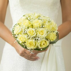 Beneath the gentle glow of northwest London's afternoon light, this bridal bouquet reflects the fusion of tradition and modern Brent Cross style. Plump, pale yellow roses nestle closely together, forming a harmonious, domed core, their perfectly unfurled petals soft as silk and fragrant with fresh promise. Delicate white sprigs of baby's breath intersperse throughout, their airy clusters bringing a sense of lightness and a dash of vintage charm-a familiar touch for weddings celebrated near Brent Reservoir or Hendon Way. Encircling the blooms, slender strands of deep green foliage or glossy decorative wire loop artfully, lending the bouquet a sculptural grace and contemporary edge. The bride's hands, expertly manicured in a soft blush polish, cradle the arrangement with affectionate reverence, the gold ring on her left hand catching just a gleam of light. The background-a subtly textured wedding gown in classic white-forms a hushed, elegant canvas that lets the bouquet take centre stage. This floral arrangement, composed with passion and expertise by local Brent Cross florists, is ideal for couples desiring a blend of old-world floral romance and the polished sophistication of a North West London celebration.