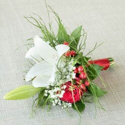 Positioned on a softly textured linen surface reminiscent of Brent Cross's boutique decor, this stunning bridesmaid bouquet captures the spirit of contemporary North London celebration. Its centerpiece is a radiant white lily, boldly unfurled, each petal pristine and slightly speckled within, cradling golden-yellow anthers that softly glow in natural light. Nestled nearby, a sumptuous red rose displays velvety layers, the spiraling petals inviting admiration-its richness a symbol of heartfelt moments shared from Brent Cross Shopping Centre to West Hendon. Delicate baby's breath, airy and pure, weaves an ethereal element through the bouquet, while clusters of glossy, cherry-red berries add unexpected festivity and cheerful contrast. Surrounding foliage ranges from delicate, feathery greens to glossy, broad leaves, evoking the lush gardens tucked away in Brent Cross neighbourhoods. A single green lily bud gently arcs from the cluster, an emblem of renewal and hope. Tied together with a gleaming satin red ribbon, each stem is secured with care and affection. This bouquet, a favourite among wedding florists in Brent Cross, makes for an elegant yet spirited statement, carrying a harmony of colours and textures ideal for memorable local celebrations and lasting impressions.