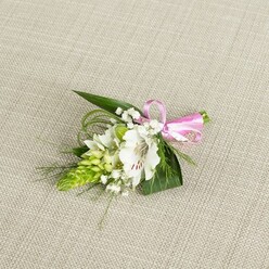 An elegant floral buttonhole is carefully arranged on a pale beige, linen-like fabric, the subtle weave of the cloth forming a calm, understated stage for the design. At the heart of the piece, a single white alstroemeria opens fully, its petals slightly ruffled at the edges and dusted with fine reddish-brown streaks near the centre, suggesting both delicacy and quiet confidence. To its left rises a compact lime-green spike of tightly clustered buds, comparable to an Ornithogalum stem, each bud forming a tiny, pointed segment that lends structure and modern flair to the arrangement. Threaded between these main elements are sprigs of airy white baby's breath, their minuscule, cloud-like blossoms softening every line and creating a gentle, romantic haze. The flowers are supported by a base of deep green foliage: broad, pointed leaves lay flat and secure, while thinner, grass-like blades curve and loop outward, adding texture and a sense of organic movement that feels reminiscent of strolling through the green corners around Brent Cross and nearby Hendon. On the right, a smooth pink satin ribbon gathers the stems together, tied into a neat, cheerful bow that introduces a touch of celebration and hints at weddings, proms, or special evenings out. The lighting is even and natural, illuminating the contrast between the crisp white petals, fresh green tones, and the soft neutral background, without any glaring shine. Every detail-down to the gentle shadows beneath the foliage and the slight sheen on the ribbon-speaks of a florist's practiced hand and of a boutonniere ready to accompany a meaningful moment in Brent Cross, whether pinned to a groom's lapel or worn for a milestone celebration.