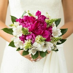 In this detailed wedding close-up, the camera focuses tightly on a bride's bouquet held against the bodice of her white gown, offering a richly textured study in magenta, white, and green. At the centre, large fuchsia peonies dominate, their petals billowing and layered like soft silk, creating a lush, cushiony core that feels indulgent and romantic. Nestled among these voluptuous blooms are white alstroemeria, each flower with slim, pointed petals and fine maroon speckles inside the throat, giving the bouquet an intricate, almost hand-painted quality. Between the main flowers, narrow, pointed green buds and spiky fillers rise up, their bright, youthful green adding energy and a contemporary edge, breaking up the softness with just the right amount of structure. A frame of deep green, slightly curved leaves encircles the bouquet, their smooth, glossy surfaces catching the light and grounding the design so that the vibrant colours appear even more vivid. The stems are gathered tightly and wrapped, disappearing into the bride's hands, where pale pink nail polish and relaxed fingers speak of calm happiness rather than nerves. Behind everything, the fabric of the dress-possibly lace with a subtle pattern-fades into a gentle blur of white, ensuring that the flowers stay in crisp, sharp focus. The image feels like a moment stolen just outside a Brent Cross venue, perhaps on a quiet side street off the North Circular, as the couple pauses with their photographer. It communicates the craftsmanship of a local florist, the freshness of early summer blooms, and the emotional weight of carrying something beautiful and carefully chosen down the aisle.
