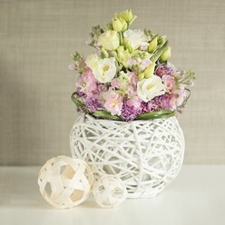 Nestled elegantly on a luminous white table, this handcrafted bouquet radiates tranquility and understated luxury, as seen through the eyes of a Brent Cross florist who delights in bringing the suburb's refined sensibility indoors. Creamy lisianthus and soft yellow eustoma, their petals opening like whispered secrets, mingle effortlessly with plush pink stocks whose vertical lines evoke the gentle rise and fall of Henlys Corner's skyline. At the arrangement's base, clusters of rich purple filler flowers - possibly alliums or asters - create a lively, textured tapestry, anchored by a soothing ring of deep green leaves. The entire floral display rises from an eye-catching spherical vessel, its open-work weave of white rattan or fine branches imparting a sense of modern artistry and organic lightness that complements contemporary North London interiors. Flanking the vase, a duo of decorative spheres crafted from pale natural wicker continue the tactile narrative, lending the scene a collected, harmonious feel. The tableau is set before a luxuriously subtle greige wall covering that softens and elevates the colors and contours of the flowers, bathing them in the day's gentle, filtered light. Whether intended for a wedding reception along High Road or as a seasonal table highlight for local gatherings, the arrangement's blend of shapes, hues, and textures reflects the attentive skill and artistic spirit that Brent Cross Flowers brings to each composition.