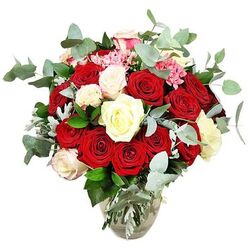 A lush and romantic rose bouquet is arranged in a clear glass vase, set against an immaculate white background that allows every hue and texture to stand out. The composition is dominated by numerous deep red and vivid scarlet roses, their velvety petals curling inwards and outwards in layered spirals, some still in tight buds, others fully open and expansive. Between these passionate reds, clusters of creamy white and soft ivory roses appear, a few tipped with a barely there pale pink, giving the whole piece a softer, more tender character. Delicate bursts of bright pink filler flowers peek through the gaps, adding a playful contrast in both colour and shape, their smaller petals giving a fine, lace-like texture that sits beautifully beside the smooth rose blooms. The bouquet is woven with plentiful blue-green eucalyptus, whose characteristic rounded, slightly frosted leaves frame the roses and suggest a cool, invigorating scent, like fresh air drifting over the greenery around Brent Cross shopping centre early in the morning. Dark green foliage and patches of silvery-green leaves round out the design, filling it to a generous, rounded silhouette that feels opulent yet thoughtfully controlled. The transparent vase, with its softly rounded base and narrower neck, reveals the stems arranged neatly in clear water, giving a sense of freshness and careful craftsmanship. The pure white background strips away any distractions, making the reds appear deeper, the whites brighter, and each curve of petal and leaf more striking. This bouquet feels perfectly suited for a declaration of love, an anniversary surprise, or refined home décor in the Brent Cross area, with same-day delivery bringing that emotion straight to the doorstep.