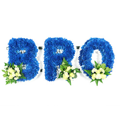 This studio portrait presents a three-part memorial spelled BRO, each letter constructed from thousands of tiny, royal blue chrysanthemum florets packed so densely they read as a soft, textured carpet of blue. Viewing at eye level, you notice the sculpted edges and the slender white foam border that helps hold each letter's silhouette-white visible in the inner loops of the B and R and across the central hole of the O-giving the arrangement a crisp, architectural clarity against the stark white backdrop. Thoughtful accenting grounds each character: a small cluster at the B's lower-left and matching clusters at the lower-right of the R and O, composed of creamy yellow or pale ivory spray roses in mixed stages of bloom, interspersed with tiny white filler blooms and sprigs of mixed greenery. The foliage choices-broader, glossy leaves reminiscent of salal or lemon leaf and feathery fern fronds-introduce tonal contrast and a faint herbaceous scent that offsets the gentle perfume of the roses. The colour palette of saturated royal blue offset by soft creams and whites yields a solemn, peaceful presence suited to a funeral chapel or wake. The arrangement reads as both personal and formal, a bespoke tribute designed to say more than words. A florist familiar with Brent Cross and nearby Golders Green would select these elements for a family seeking a dignified, clear statement of remembrance at a memorial service, where the blue symbolises calm and the pale roses offer consolation.