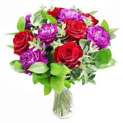 This bouquet brings together the drama of red roses and the softness of purple peonies in a way that feels perfectly at home in Brent Cross, as if styled for a special dinner party just off Hendon Way. The roses are the first thing your eye finds: large, velvety blooms, fully opened to reveal spirals of deep crimson petals that look almost plush to the touch. Nestled alongside them, the peonies offer a contrasting form, their blossoms rounder and more expansive, with layers of ruffled purple petals that suggest abundance and quiet luxury. Encircling these blooms is a generous frame of fresh greenery. Smooth, rich green leaves create a sense of fullness and vitality, while long, elegant stems of eucalyptus thread through the bouquet, their subdued silvery tones softening the bold colours and adding a hint of airiness to the composition. The flowers are gathered into a clear glass vase, where you can see the stems arranged with precision, each one cleanly cut and fanned out in the water. The glass itself reflects small flecks of light, perhaps similar to the gentle glow of evening lights around Brent Cross. Soft, natural light seems to wash over the bouquet, enhancing the reds so they almost smoulder and making the purples appear deeper and more intriguing. You can almost sense the mingling scents: the warm, familiar fragrance of roses, the faint, green freshness of cut leaves, and the crisp lift of eucalyptus. This is the sort of arrangement that suits meaningful occasions-a romantic gesture sent through Brent Cross Flowers, a congratulations bouquet for a promotion, or an uplifting gift to brighten a favourite corner at home.