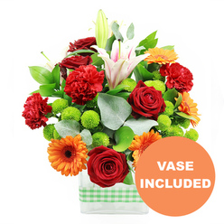 This photograph presents a vibrant, carefully structured bouquet that feels perfectly suited to a cheerful living room near Brent Cross station or a bright kitchen by the North Circular. The arrangement is housed in a modern rectangular glass vase, clear and minimalist, allowing the viewer to see the tidy stems inside. Wrapped around the base is a light green and white gingham ribbon, its soft checked pattern providing a touch of homeliness, like something you might see on a family table after a weekend visit to Brent Cross Shopping Centre. Center stage in the bouquet are several deep red roses, their lush, velvety petals curled inward in dense spirals, radiating warmth and a classic romantic note. Around them, red-orange carnations spread out with ruffled petals, adding a soft, cloud-like texture that offsets the bold structure of the roses. Cheerful orange gerbera daisies appear throughout, each bloom sporting a ring of smooth, flat petals around a dark centre, bringing a sunny, almost playful quality. Tall white Oriental lilies rise gracefully above, some fully opened with visible stamens and delicate pink striping along their inner petals, others still in bud form, suggesting days of continued blooming. Tucked between these focal flowers are clusters of small, round lime-green pompom chrysanthemums, their spherical shapes and fresh colour lending a bright, zesty highlight that keeps the arrangement lively. Lush green foliage frames everything: there are broad deep-green leaves and softer, silver-green eucalyptus stems that trail and curve, adding dimension and a more natural garden feel, as if freshly arranged by a local Brent Cross florist for same-day delivery. The image is set against a pure white backdrop, emphasizing the bouquet's colourful contrasts. In the lower right corner, an orange circular graphic overlay clearly states "VASE INCLUDED" in bold white letters, signalling that the sleek glass vase seen here arrives as part of the finished gift.