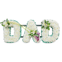 In this detailed funeral arrangement, the word DAD is shaped from three robust block letters, each one completely carpeted in immaculate white chrysanthemums or similar small white blooms. The flowers are tightly massed so no stems or gaps can be seen, giving the letters a thick, plush covering that appears soft to the touch, like a peaceful bed of petals. The uniform whiteness conveys purity, serenity, and a quiet sense of farewell. Encircling the edges of every letter is a vibrant emerald green satin ribbon, gathered into gentle ruffles. This ribbon forms a clean, defined border, the rich green hue contrasting against the white flowers and suggesting renewal and the calm of nature. Behind the letters, faint horizontal bars of a dark frame can be glimpsed, indicating that the tribute is structured to stand upright during a service in Brent Cross, perhaps placed near the front of a synagogue, church, or crematorium close to Brent Cross station.   

Delicate accent flowers add movement and emotional nuance. On the upper left side of the first D, a small spray of white freesias and glossy green foliage rests softly atop the chrysanthemums. From this point, two thin, arching green stems sweep diagonally towards the central A, visually linking the letters. At their endpoint, a striking cluster forms the arrangement's focal point: two deep purple calla lilies, with their sculptural, trumpet-like blooms, are nestled among smaller white freesia and fresh greenery, introducing a rich, dignified contrast. The last D bears a more compact version of this detail, with white freesia and foliage accenting its upper right side. Altogether, the piece feels personal and composed, like the kind of bespoke DAD tribute a Brent Cross family might order to express love, gratitude, and cherished memories of a father who anchored their home.
