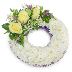 Against a clean white field sits a carefully composed circular funeral wreath, the sort of tribute you might see brought from a Brent Cross florist to a local church or cemetery near Cricklewood. The majority of the design is formed from tightly packed white spray chrysanthemums-the small, ruffled heads overlapping like petals on a snowy tide-producing a richly textured, consoling surface that feels both substantial and delicate. Thin flashes of a deep purple netting or ribbon trace the inner and outer edges, glinting discreetly through the massed white blooms and lending the wreath a quiet, elegant outline. Nestled on the upper-left arc, three soft yellow roses open with gentle dignity, their creamy tones evoking friendship and consolation; around them float clouds of baby's breath, lending an ethereal, lace-like softness, while small clusters of lavender-purple statice supply a muted, complementary counterpoint. Varied greens-broad, smooth leaves and feathery fern fronds-embrace the focal spray, their tactile contrast amplifying the lushness of the flowers. The photograph's soft, natural light highlights the petals' textures and the gentle shadow beneath, suggesting late morning or calm studio illumination. Sensory impressions come through: the cool silk of chrysanthemum petals, the faint warm perfume of roses, and the dry, slightly herbal whisper of statice. Intended for a dignified farewell in the Brent Cross area, this wreath is designed to convey sympathy, steady support and a graceful sense of continuity during a time of parting.