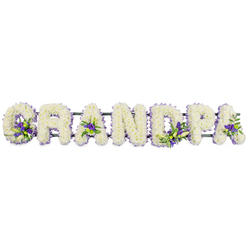 A lovingly constructed funeral flower arrangement spells out GRANDPA in bold, upright capital letters, laid out horizontally as though placed at the front of a chapel or crematorium in the Brent Cross area. Each letter is densely covered with pristine white chrysanthemums or carnations, the small blooms set closely together to create a cushion-like depth and a velvety surface that feels soft even at a distance. The white flowers form a calm, unbroken canvas that speaks of purity, reverence, and quiet reflection. Resting upon this blanket of white are measured accents of purple and lavender flowers, perhaps lisianthus or petite irises, combined with sprigs of fresh green foliage. These contrasting clusters are not overdone; instead, they appear at thoughtful intervals-the lower curve of the G, the inner triangles of the As, the arc of the D, and the bowl of the P-adding just enough colour to suggest affection and memory without overwhelming the overall serenity. A delicate violet ribbon follows the outline of each letter, framing the shapes and helping them stand out clearly, similar to how a nameplate might be highlighted on a memorial. Beneath the blossoms, a discreet dark green framework links all the letters into one stable design, making it practical to transport from Brent Cross Flowers to homes, chapels, or cemeteries around Golders Green and Cricklewood. The composition is both visually striking and emotionally gentle, offering a personalised floral tribute that families might choose when they wish to say goodbye to a grandfather with something that feels intimate, carefully made, and deeply respectful.