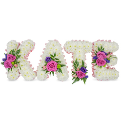 Presented against a clean white studio background, the name KATE appears in four freestanding letters, each letter built from thousands of tightly packed, snow-white chrysanthemums that form a plush, tactile surface you could almost trace with a fingertip. A gentle blush of pastel pink satin ribbon outlines each character, softening the crisp white and catching the light with a faint, satiny glow. On top of that pure base, vivid clusters of velvety fuchsia-pink roses and delicate pastel pink spray roses emerge like warm, affectionate notes; pockets of deep purple or blue blooms-probably statice or small lisianthus-add an elegant, cool punctuation. Greenery threads through the design-feathery ferns, glossy eucalyptus leaves, and fine-leaved fillers-creating pockets of shadow and depth so the letters read as sculpted forms rather than flat panels. The floral accents are deliberately placed: the K carries a bold cluster along its diagonal stroke, the A is softened by blooms at its lower right curve, the T has emphasis near its base, and the E is balanced by flower groupings on the middle bar and lower arm, guiding the eye across the name. The arrangement gives off a subtle rose perfume over crisp chrysanthemum freshness, evoking gentle consolation. As a sympathy or memorial tribute, this composition feels intimate and composed-appropriate for a chapel service, graveside remembrance, or a quiet farewell in the Brent Cross and Cricklewood area-crafted with the steady hand of a local florist who understands how flowers carry memory.