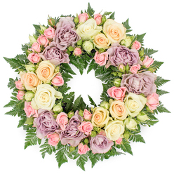 The image presents a full, circular wreath designed as a funeral or memorial tribute, positioned centrally against a crisp white background so that its colours and textures show clearly. The wreath itself is a continuous ring of roses, tightly massed together to create an opulent, almost cushion-like surface of petals. Soft ivory and creamy white roses dominate, many of them fully open, revealing the intricate swirl of their inner petals and catching subtle highlights that give the design a gentle radiance. Interspersed among these are roses in understated hues of pale peach and buttery yellow, which lend a warm undertone and soften the overall appearance. Smaller blush pink spray roses nestle between the larger heads, appearing in tiny clusters and scattered buds that bring granularity and intimacy, as if each little flower carries a personal message of love. Dusty lavender and muted mauve roses are carefully placed around the ring, their antique shades bringing depth and a note of quiet reflection. Underpinning the entire composition, rich, dark green fern fronds extend outward and peek between blooms, their feathery texture contrasting with the smoother rose petals and shaping the wreath's perfectly round outline. The balance of colours and forms suggests the work of an experienced Brent Cross florist, attuned to creating designs that feel both classic and heartfelt for local families. The wreath feels peaceful and contemplative, well suited to funerals, cremation services, or memorials in and around Brent Cross, where its soft palette and elegant composition would offer a calming focal point during moments of farewell.