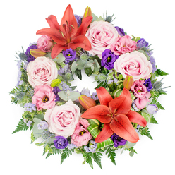 This elegant sympathy wreath is shown from above against a pure white backdrop, its circular shape a gentle reminder of continuity and eternal love. The base is a luxuriant ring of mixed greenery, built from silvery eucalyptus-like leaves, layered fern fronds, and other deep green foliage that together create a soft yet substantial structure. Nestled into this verdant frame are two bold coral-orange Asiatic lilies, each fully open with wide, star-shaped petals and clearly visible stamens, one placed toward the upper curve of the wreath and the other slightly off-centre below to create a balanced focal rhythm. Surrounding and softening these vivid lilies are numerous roses in shades of blush and lavender-pink, their petals arranged in intricate spirals that feel both delicate and reassuring. Interwoven among the roses, ruffled eustoma (lisianthus) blossoms appear in pale pink and rich, velvety purple, their cup-shaped blooms adding little pools of shadow and depth, inviting the eye to pause and reflect. Standing proud above the softer forms, small blue-grey eryngium thistles add a cool, architectural accent, while fine sprays of tiny lavender-blue filler flowers drift through the design like a light mist, uniting the colours with gentle cohesion. A few partially closed lily buds are tucked discreetly among the greens, suggesting that not every story has finished, even in moments of farewell. Near the lower lily, bright green leaves have been folded and arranged with a sculptural flair, showcasing skilled floristry that would be at home in any Brent Cross ceremony or gathering. The overall impression is one of quiet dignity and tender emotion, a wreath suitable for funerals or memorials across Brent Cross and the surrounding North West London neighbourhoods, offering comfort through its thoughtful composition.