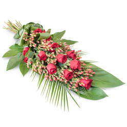 A refined sympathy spray of red roses, berries, and lush foliage lies diagonally across a bright white background, evoking the peaceful stillness of a farewell gathering in Brent Cross. The arrangement is long and slightly tapered, with the stems gathered at the top left and the flowers and greenery fanning gracefully towards the lower right. Along its length runs a continuous line of velvety red roses, some still modestly closed, others opening to reveal intricate spirals of soft petals that seem almost touchable. Threaded between these blooms are clusters of round, pinkish-red Hypericum berries whose small, polished forms bring a jewel-like glimmer and subtle variation in tone. The base of the spray is built from sturdy, glossy dark green leaves laid in overlapping layers, forming a rich, leafy foundation that anchors the vivid reds and gives the piece a sense of depth and comfort. From the lower right corner, delicate palm fronds reach outward in airy strokes, extending the silhouette and adding a gentle sense of movement, as if a soft breeze had passed through. At the top left, the stems are tightly grouped and wrapped with a folded green leaf band, creating a natural, tidy finish that reflects the craftsmanship of an experienced local florist. The stark white background strips away distraction, allowing the interplay of red petals, green foliage, and berry texture to tell a quiet story of remembrance. This classic funeral spray would look entirely at home resting near a chapel altar, by a crematorium service in the Brent Cross or Golders Green area, or accompanying a final farewell at a local cemetery.