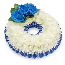 Captured from a gentle overhead angle on a stark white surface, this circular sympathy wreath presents a composed study in texture and tone that a local florist might bring to a memorial near Brent Cross Shopping Centre or Staples Corner. The foundation is an abundant band of pure white chrysanthemums, hundreds of tiny petals building a plush, slightly domed ring whose soft surfaces catch the light and reveal the blooms' fine, feathery detail and the faint, warm hint of cream at their centers. Anchoring the design on the upper left are three vivid royal-blue roses - petals layered in a tight, velvet spiral whose colour reads almost electric against the white field - surrounded by glossy deep-green foliage that frames and supports the focal cluster. A pleated satin ribbon in the same deep cobalt blue trims both the outer and inner rims of the wreath; its slight luster and ruffled texture define the silhouette and bring an ordered elegance to the whole piece. As an experienced local florist, I see this arrangement as deliberately restrained yet deeply personal: the white chrysanthemums speak of peace and purity, the blue roses of solace and steadfast memory. The composition's soft shadow and balanced proportions give it a quiet presence suitable for a funeral reception or chapel display, and Brent Cross Flowers prepares similar tributes with attention to texture, proportion, and the gentle symbolism families seek when laying a loved one to rest.