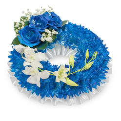 A thoughtfully composed memorial wreath that reads like a quiet hymn in blue and white: a dense ring of small, vibrant deep-blue chrysanthemums creates a tactile, cushiony base that dominates the circular form. Their intricate, needle-like petals capture a soft, muted glow, producing a textured field of sapphire that feels steady and consoling. Lining the inner circle, a ruffled white satin ribbon gives the piece an elegant frame; its glossy folds reflect a cool light and add a refined softness against the intense blue. Nestled at the upper-left quadrant, three large blue roses bloom fully-petals thick and velvety, unfolding in a rich azure that complements the chrysanthemum backdrop. Delicate clouds of Gypsophila (baby's breath) surround the roses, lending a sense of air and delicate contrast, while dark green foliage grounds the cluster. A graceful cascade of white orchids arcs from mid-left toward the lower right: their smooth, porcelain-like petals and subtle, floral scent introduce an airy serenity as they float above the dense blue. Among them, fresh green orchid buds promise growth and continuity, some just beginning to open. The composition balances texture-the spiky chrysanthemum heads, velvety roses, silky orchids-and mood, moving from solemn remembrance to quiet hope. Picture this arrangement laid near a lectern in a chapel on a cool, overcast afternoon, or placed by family at a graveside service in and around Brent Cross and nearby Hendon; its restrained palette and careful craftsmanship make it a dignified expression of sympathy and enduring respect.