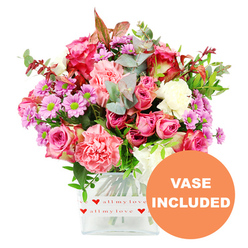 This photograph shows a generous, romantic bouquet arranged in a clear, square glass vase and set against a spotless white backdrop, so the floral colours appear especially vivid. At the core of the design are several large pink roses, some with deep fuchsia tones and others a softer, more pastel pink, their layered petals unfolding in a classic, elegant spiral. Around them, plentiful carnations in coral, blush, and creamy white form a cushion of ruffled texture, creating a sense of softness you can almost feel under your fingertips. Scattered throughout are small daisy-like chrysanthemums in shades of lavender and purple, each bloom finished with a bright green centre that punctuates the arrangement with cheerful, eye-catching dots. The flowers are surrounded by a halo of fresh greenery, including silvery-green eucalyptus leaves and darker, glossy foliage, with just a few reddish-brown accents adding warmth and depth, like the colours seen in nearby Brent Cross parks at the turn of the season. All the stems are tidily gathered into a modern glass vase whose base is wrapped in a white band decorated with little red hearts and the handwritten-style phrase all my love, transforming the arrangement into an unmistakably affectionate gesture. In the lower right corner of the image sits an orange circular graphic, stamped with the words VASE INCLUDED in bold white capitals, clearly indicating this bouquet arrives complete and ready to display. The mood is one of thoughtful generosity and fresh local floristry, ideal for same-day delivery from Brent Cross Flowers to loved ones in nearby Hendon, Cricklewood, or along the Edgware Road when you want to brighten a room and a day in one beautiful moment.