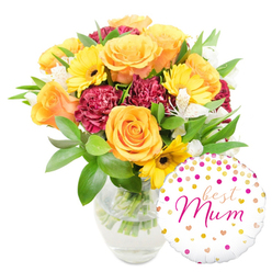 Set against a clean white backdrop, this uplifting gift pairing shows a lush bouquet arranged slightly to the left in a transparent glass vase and a cheerful round foil balloon positioned to the bottom right. The bouquet glows with honeyed warmth: peachy-orange and golden yellow roses form the heart of the arrangement, their velvety petals unfurling in soft whorls. Scattered among them, sunny yellow gerbera daisies offer bright, open faces with deep, cocoa-coloured centres that draw the eye. Rich burgundy carnations, densely ruffled, bring a romantic, tactile weight, while small white alstroemeria with their delicate, speckled throats weave a subtle, elegant contrast. Abundant glossy greens - from broad leaves to feathery sprigs - cradle the blooms and create a natural, garden-fresh silhouette, and the clear vase exposes neatly trimmed stems in fresh water, a visual cue of recent, careful floristry.

The balloon, glossy white and dotted with gold and vivid pink confetti-like spots and tiny hearts, carries the affectionate inscription best Mum in a lively pink script, overlapping the vase in a playful, personal way. Soft natural light bathes the scene, highlighting petals' textures and the reflective glass without harshness, and the composition has a relaxed, joyful energy that feels like a sunny kitchen table on a morning visit. You can almost sense the heady, slightly sweet scent of roses mingling with green cut stems. Thoughtfully composed for Mother's Day, a special birthday or a 'just because' surprise, this bouquet is the kind of arrangement frequently requested by families in Brent Cross and neighbouring Cricklewood, offering warmth, freshness and a reassuringly local touch.