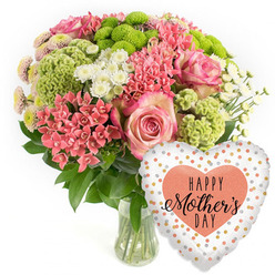 A lush Mother's Day floral gift is captured in a bright, uncluttered image, making every bloom, leaf and reflection stand out. In the centre sits a clear glass vase, filled with clean water and carefully trimmed stems, supporting a rounded bouquet that feels both full and airy. Prominent within the design are large pink roses with gently curving petals, the centres rich and rosy, the outer edges softly fading to paler shades, like a gentle blush. Around the roses, dense clusters of coral-pink flowers-evoking kalanchoe or similar small florets-create texture and warmth, while the occasional spikier coral bloom adds a touch of spirited movement, echoing the swirl of people and traffic around Brent Cross. Lime-green button chrysanthemums and plush, chartreuse, cockscomb-style forms pop from the arrangement, their vivid colour adding an invigorating jolt and their unusual, almost moss-like surfaces making the display visually intriguing. Scattered white spray chrysanthemums, with bright daisy-like faces, introduce pockets of lightness, ensuring the palette remains crisp and uplifting. Fine details, such as tiny pale-tipped chrysanthemum centres and overlapping dark green leaves, reward a closer look, revealing the florist's care and craftsmanship, much like the attention to detail you'd find at Brent Cross Flowers. The bouquet is photographed from slightly above and to the side, showing its domed shape and the pleasant balance of colour. To the right of the vase, a reflective heart-shaped foil balloon brings a festive flourish. Its white background is decorated with irregular, shimmering dots of gold, silver and soft coral-peach, and in the middle a solid peach heart frames the message "HAPPY Mother's DAY" in elegant black letters. Together, the arrangement and balloon radiate affection and appreciation, ideal for a Mother's Day delivery to homes and apartments across Brent Cross.