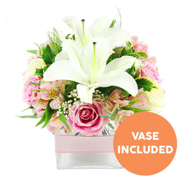 This image presents a carefully composed bouquet set against a pristine white background, the simplicity of the setting allowing every flower to stand out as if lit by soft daylight filtering into a Brent Cross apartment. At the centre of the picture, two fully open white Oriental lilies form the main focal point, their sculptural petals gently arching back to frame clusters of bold reddish-brown stamens and central pistils. Encircling the lilies, soft pink roses form a rounded collar, with petals in various stages-some still tightly wound in a classic spiral and others relaxed and open, revealing layer upon layer of smooth, velvety texture that speaks of romance and gentle celebration. Interspersed among the larger blooms are stems of pink and white alstroemeria, their elegant petals slightly flared, several lightly speckled with yellow, adding subtle pattern and interest to the design. Wisps of delicate white baby's breath weave through the bouquet like tiny stars, lending a sense of lightness and filling the spaces without overwhelming the central flowers. The arrangement is backed by rich greenery, including broad, shiny leaves that reflect the light and feathery fronds that introduce a sense of movement, similar to trees swaying along the paths near Brent Cross Park. All the stems converge neatly in a low, square clear glass vase, the waterline visible and the green stems clearly seen, giving the design a fresh, contemporary feel. Around the middle of the vase, a pale pink satin ribbon is wrapped smoothly, adding a soft decorative accent. In the lower right corner of the image, an orange circular badge with bold white capital text reads "VASE INCLUDED," clearly highlighting that the vase forms part of the floral gift, ideal for same-day flower delivery to homes and offices around Tilling Road and the North Circular.