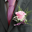 This image focuses tightly on the chest and shoulders of a person dressed for a formal occasion in Brent Cross, perhaps a groom waiting outside a local registry office or a guest stepping out of a car near the shopping centre's event spaces. The suit jacket is a deep charcoal, almost black, with the left lapel crisp and defined in the frame, its texture smooth yet substantial. Beneath it sits a light-coloured dress shirt, patterned with a fine gingham or check that adds a quiet sophistication. Down the centre runs a vivid magenta-pink tie, impeccably knotted and glossy, providing a confident splash of colour that draws the eye immediately. Anchored to the lapel is a carefully arranged boutonniere: a single pale pink rose in full bloom, its softly curling petals fading from a richer pink at the centre to a tender blush along the edges, suggesting both youthfulness and tenderness. Encircling the rose are airy clusters of white baby's breath, their tiny, star-like blossoms forming a gentle, clouded border that softens the strong lines of the suit. Behind the central bloom, slender blades of fresh green foliage rise and fan out lightly, introducing a sense of living movement, as if touched by a light spring breeze over nearby Clitterhouse Playing Fields. The base of the floral piece is wrapped neatly in a deep green leaf or ribbon, cinched with a satin bow in muted purple or mauve. The bow's sheen echoes the tie's lustre, creating a harmonious and contemporary colour story. The overall impression is one of poised celebration and affectionate detail, capturing the way a thoughtfully chosen buttonhole can add personal meaning and romantic symbolism to formal wear at Brent Cross weddings, proms, or gala evenings.