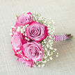 Against a pale beige fabric that looks softly woven and lightly textured, a small, elegant bouquet is placed on a diagonal, as if resting briefly between photos at a Brent Cross wedding. The heart of the arrangement features several open roses with striking two-tone petals. Each bloom begins with a cool lavender or light purple centre and gradually transitions into a deep, saturated fuchsia and magenta pink along the outer petal edges, creating a bold yet refined contrast. These roses appear full and velvety, their layers of petals curling outward in gentle spirals that catch shadows and highlights. Interwoven with the roses are generous sprays of white baby's breath, whose tiny, numerous blossoms form airy clusters that soften the vivid colour of the petals. The gypsophila adds an ethereal, cloud-like texture, filling spaces between the larger blooms and lending the bouquet a sense of lightness and innocence, much like the fresh air you might feel walking from Brent Cross station to nearby parks. The stems are carefully aligned and tightly bound with a deep purple satin ribbon, smooth and slightly glossy, creating a strong line that contrasts with the softness above. Wrapped around this ribbon, a string of white faux pearls coils in a graceful spiral from top to bottom, giving the handle a decorative, bridal finish that feels both classic and contemporary. With the pearl-wrapped handle pointing towards the right, the bouquet looks poised to be carried down an aisle, offered as a prom posy, or used as a delicate accent in an engagement shoot around Brent Cross and Hendon. The mood is intimate, romantic, and thoughtfully styled, showcasing the artistry of a local florist who understands the area's blend of modern style and timeless tradition.