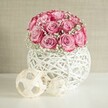 On a smooth white tabletop, a rounded bouquet of soft pink roses and airy white baby's breath sits in a sculptural, white woven vase, creating a calm, romantic still life that feels perfectly at home in a modern Brent Cross apartment. Around fifteen to twenty roses form a lush dome, each bloom opening in layered spirals. Their petals fade from a pale blush at the heart to a richer, almost fuchsia pink along the ruffled edges, giving the flowers a gentle ombré effect that suggests warmth and tenderness. Between the roses, tiny clouds of gypsophila float, their miniature white blossoms softening the pink tones and adding a sense of lightness, like mist caught in morning light over nearby Clitterhouse Playing Fields. The flowers appear fresh and velvety, as if they have just arrived from a local florist, their scent imagined as sweet but not overpowering. They are cradled in a spherical white container made from interlaced strands that resemble painted rattan or wicker, the open criss-cross pattern allowing glimpses of shadow and reflected light to dance through. To the left of the vase, two decorative woven spheres in pale beige echo the natural textures, their thin wooden strips looping and crossing in loose, geometric patterns, one noticeably larger than the other. Behind the arrangement, a softly textured wall in warm beige or off-white creates a serene backdrop, with diffused, even lighting that casts gentle, almost imperceptible shadows. The overall composition feels tranquil and polished, ideal for a coffee table scene in a Brent Cross home or as inspiration for an intimate, handcrafted centerpiece for anniversaries, birthdays, or a quiet evening in.