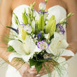 This wedding bouquet reads like a careful promise: large, star-shaped white lilies open generously, their velvety petals reflecting soft daylight, while purple-tinged lisianthus provide a refined, painterly contrast that deepens the palette without stealing warmth. Tucked among these main blooms are tiny veronica spikes and slender buds that add whisper-fine height, and sprigs of baby's breath that work like miniature clouds between the more sculptural shapes. Fine, grassy stems and delicate filigree foliage weave through the arrangement, creating an airy, moving silhouette that makes the bouquet feel both substantial and effortless to hold. The scent is a gentle, floral sweetness with crisp green undertones-an aroma that suggests dew-damp mornings in a neighbourhood garden, familiar to brides who pick Brent Cross or nearby Hendon for their photographs. Daylight in the photo is soft and forgiving, enhancing the creamy whites and cool violets while allowing shadows to reveal petal texture and depth. As a florist in the area, I assembled each stem with attention to balance and rhythm so every blossom-open or in bud-contributes to a cohesive, heartfelt story. This piece would sit naturally in a range of settings, from a traditional church near Golders Green to a contemporary ceremony by the North Circular, carrying an air of quiet celebration and considered luxury typical of local wedding arrangements from Brent Cross Flowers.
