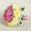 On a softly woven, neutral fabric surface in warm beige tones, this beautifully composed hand-tied bouquet is arranged as though waiting for a bride in a Brent Cross hotel suite or family home. Seen from a slightly raised viewpoint, the bouquet's round shape is perfectly balanced, its blooms forming a harmonious circle divided into two complementary halves. One side features vivid fuchsia and magenta-pink roses, each head fully open, revealing a tight spiral of velvety petals that suggest depth and richness. The opposing side is fashioned from creamy, pale yellow roses, their petals broader and softly layered, giving a gentle, luminous quality that contrasts elegantly with the stronger pinks. Throughout the entire design, dainty white baby's breath is threaded like a fine veil, its tiny blossoms clustering in between the roses, adding softness and a delicate, traditional charm. Tucked discreetly among the flowers, deep green leaves offer structure and a touch of garden freshness, reminiscent of walks through nearby parks in Hendon or Childs Hill. The stems are drawn together into a neat, cylindrical handle, expertly wrapped in smooth pink satin ribbon with a subtle sheen. Winding around this ribbon, a single row of white pearls trails in a graceful spiral, lending the bouquet an air of refined luxury, as if it were an heirloom accessory. The arrangement feels poised and serene, perfectly suited for wedding ceremonies around Brent Cross, anniversary celebrations, or romantic occasions where elegance, sentiment, and the timeless symbolism of pink and yellow roses are cherished.