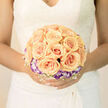 This arrangement has the softness of a remembered morning: a symmetrical cluster of apricot roses at its core, each bloom unfurling like a tiny sun caught mid-dawn, their creamy outer petals blushed with warm peach. A ring of delicate lisianthus-white centres shaded to pale violet at the edges-frames the roses, providing a cool, twilight-like punctuation that balances the bouquet's glow. Interspersed among them, delicate sprays of white baby's breath and other airy fillers create a light, drifting lattice that makes the composition feel weightless, as though it might lift on a warm breeze. The lighting in the image is tender and even, blurring hard edges and softening contrasts so the whole arrangement reads as a gentle, dreamlike object; shadows are subtle and there are hints of translucence where sunlight skims the petal margins. The textures shift from the rich, creamy density of the rose centers to the crisp, feathery accents of the filler blooms, producing an overall sensation of refined delicacy. This bouquet is the kind brides choose when they want something that reads as romantic without being ornate-timeless in shape, intimate in scale. I often create variations like this for ceremonies and portraits at local venues around Brent Cross and Hendon, where understated elegance suits the light and architecture; it carries the quiet assurance of a well-loved tradition, meant to be held close and remembered long after the vows are spoken.