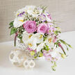 A refined floral basket arrangement sits on a smooth, reflective white surface, radiating the soft romance of an elegant Brent Cross wedding. The white wicker basket, intricately cross-hatched and neatly finished, cradles an abundance of blooms that tumble gracefully over its edges in a gentle, asymmetrical cascade. At the centre, generous pink roses are fully open, their plush, spiral petals catching the light and adding a rich, velvety depth. Around them, white Phalaenopsis orchids extend outward on slender stems, their broad, porcelain-like petals and golden throats bringing a sophisticated, modern edge. White lisianthus blossoms, some tinged with a whisper of lavender at the tips, weave between the roses and orchids, accompanied by tiny stars of white baby's breath that lend an airy, cloudlike softness. Delicate clusters of smaller, blush-toned blooms, suggestive of stock or Sweet William, spill downward, further enhancing the sense of movement. A single, tall spike of deep magenta veronica rises from the basket, giving vertical rhythm and a bold note of colour that gently contrasts with the pastel palette. Layers of fresh green foliage peek through-broad leaves, tender buds, and flexible stems-creating a garden-inspired feel reminiscent of quiet walks near Brent Cross's leafy corners and residential streets. Two pale, openwork rattan spheres rest in front of the basket, echoing the natural materials and adding sculptural interest. Behind it all, a neutral beige fabric backdrop with a soft, woven texture grounds the scene, allowing the whites, pinks, and greens to glow. This arrangement feels like a tender promise, perfect for gracing a bridal table, an intimate ceremony, or a special delivery to a loved one across Brent Cross.