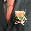 A small, artful boutonniere rests on a charcoal lapel, crafted to read like a gentle promise on a day of ceremony. The focal peach rose is immaculate, its petals layered in soft peach and creamy blush that glow against the suit's dark weave; each petal catches light like a memory held close. Surrounding it, clusters of tiny white gypsophila float in airy sprays, their delicate blooms creating an almost cloud-like halo that frames the rose without stealing its presence. Sprigs of fine green foliage - thin fern fronds and lively leaf tips - bring a whisper of the garden, introducing fresh texture and a hint of early-morning dew. At the stem the arrangement is tied with a satin ribbon in coral-pink that mirrors the tie, the bow small, neat, and hand-finished, suggesting a careful florist's touch. The overall composition is elegant and restrained, the suit's charcoal tone amplifying the rose's warmth and the white accents' purity, resulting in a boutonniere that balances romance with refinement. There is a soft, floral scent imagined in the piece, subtle enough to comfort and uplift through vows and speeches. As a Brent Cross florist I picture this pinned on a groom at a boutique venue near Brent Cross Shopping Centre or slipped into the lapel during photos along Cricklewood Broadway - a tiny, wearable heirloom. Brent Cross Flowers prepares these with attention to longevity and wearability, ideal for weddings, anniversaries, or special commemorations across the local area.
