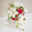 In this image, a beautifully composed bouquet captures the mood of an elegant Brent Cross wedding celebration. Large, creamy white lilies form the central height of the arrangement, their petals open and expansive, each bloom confidently facing outward as if greeting guests. Tucked around them, classic red roses add volume and intensity; their velvet-like petals spiral densely, creating a rich, romantic heart to the design. From one side, pale orchid blooms gently trail, their smaller, delicate petals providing a subtle cascade that draws the eye downward, much like the soft flow of a bridal gown as it moves through a local ceremony near Brent Cross station. Between the dominant flowers, wispy gypsophila is threaded like tiny lace, its numerous miniature white blossoms introducing a light, clouded texture that diffuses the strong red-and-white contrast. Here and there, clusters of glossy berries appear, round and polished, in a lively red-coral hue that adds a festive sparkle, reminiscent of celebratory toasts in nearby restaurants and venues. The bouquet is presented in a simple white woven vessel, whose gentle basket weave suggests craft and care without pulling focus from the blooms themselves. The composition is rounded yet airy, with each flower given space to breathe, allowing the lilies, roses, orchids, and berries to harmonise rather than compete. If you could lean closer, you might sense the heady fragrance of lilies blending with the more familiar perfume of roses, a scent that would fill a reception space or family home in Brent Cross with warmth and tenderness. This is a bouquet designed to mark life's important promises-marriage, commitment, enduring love-expressed through classic flowers arranged with modern finesse.
