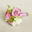 A compact, hand-tied bouquet lies horizontally on a light tan, textured fabric that looks like natural linen, the neutral backdrop gently echoing the calm, understated elegance of a Brent Cross townhouse interior. At the centre of the posy, two fully open pink roses and a third, slightly more tucked rose draw the eye, their petals unfolding in soft blush tones that deepen to a richer magenta at the very edges, creating a delicate ombré effect. The roses appear lush and velvety, each layer of petals carefully spiralled, suggesting the quiet romance of a Kensington Gardens stroll at dusk. Nestled between the roses are rounded tufts of lime-green button chrysanthemums, their tiny, tightly packed petals forming playful, cushion-like domes that bring a fresh, modern edge to the design. Pale, creamy white blossoms, likely lisianthus or freesia, weave through the composition, their lighter tones and softly ruffled petals adding airiness and a sense of movement, like a breeze drifting over Brent Cross's leafy streets. Tiny white star-shaped flowers with golden centres, reminiscent of waxflower, flicker through the bouquet, adding fine detail and a gentle, almost bridal charm. The stems are neatly aligned and trimmed, then wrapped in a bright fuchsia satin ribbon that shimmers subtly under soft, even lighting. A slender strand of white faux pearls winds diagonally over the ribbon, crossing and looping as if hand-finished by an experienced local florist, adding a hint of vintage glamour. The bouquet's overall mood feels perfect for a bridesmaid's posy, intimate registry office wedding near Brent Cross, or a delicate centrepiece for a small celebration at home, radiating freshness, tenderness, and thoughtful craftsmanship in every bloom and texture.