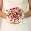 Held close against the detailed lace of a bridal dress, this bouquet feels like a captured breath of calm in the midst of wedding-day excitement, as if the bride has paused for a moment in a quiet corner near Brent Cross shopping centre. At its core, lush lavender-pink roses open generously, their plump, velvety petals forming soft whorls that invite a lingering touch. Threaded among them, zesty lime-green button chrysanthemums pop through with bright, spherical blooms, introducing a modern, almost playful note to the otherwise timeless design. Circling the edge of the bouquet, creamy-white flowers and their slender buds form a soft halo, a cloudlike frame that lets the central colours sing without ever becoming overpowering. The bouquet sits comfortably in the bride's hands, its rounded, hand-tied shape suggesting both care and ease, as though it has been gathered from a carefully tended garden just off the streets of Brent Cross and Hendon Way. Natural light highlights the interplay of tones-dusky mauve, bright spring green, and gentle ivory-while the imagined fragrance combines the familiar warmth of rose with the crisp, refreshing scent of fresh foliage. Every stem appears thoughtfully placed, chosen not only for beauty but for resilience, ensuring the bouquet remains radiant from the moment the bride steps out for photos by the local venues to the final farewell. It is an arrangement that speaks softly of joy, hope, and the comfort of love, expressed through colour, texture, and scent.
