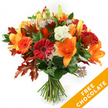 A hand-tied autumn bouquet photographed against a crisp white background, the kind of arrangement a Brent Cross florist would gather on an early, cool morning. At its heart are wide-open, trumpet-shaped orange lilies with visible stamens and a few graceful buds that promise a second wave of blooms. Scattered among them are plump Gerbera daisies in deep red and bright orange, their concentric petals forming cheerful, sun-like faces. Soft, ruffled white Lisianthus punctuate the warmth with a clean, creamy contrast, while delicate purple-pink Astrantia adds starry texture and an almost lacy intricacy to the composition. Rich, deep-green foliage-likely salal or ruscus-builds a verdant base, and most strikingly, several deeply lobed oak leaves in burgundy and burnt orange are woven through the stems, lending true autumnal structure and a hint of woodland rustle. The stems are gathered in a classic hand-tied fashion and finished with dark twine or raffia, the raw fibre giving a tactile, crafted feel. Soft, even lighting highlights petal sheen and the warm saturation of the colours, making the bouquet feel luminous yet grounded. The image also carries a bright orange diagonal banner across the lower right corner bearing the bold white text FREE CHOCOLATE, clearly part of the photograph rather than the flowers themselves. This bouquet evokes the smell of warm spice and green stems, a seasonal gift suitable for thanksgiving tables, birthdays in October, or a cozy surprise for someone returning to a home in Brent Cross or nearby Hendon. The overall impression is generous, comforting and expertly arranged by a neighbourhood florist attentive to texture and seasonal colour.