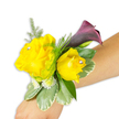 A delicate wrist corsage rests diagonally across a light-to-medium forearm against a crisp white background, its composition balanced and quietly celebratory. At the heart are two buttery yellow roses - one fully opened and sunlit to the left, the other a slightly smaller, more cupped bloom to the right - each petal soft and petaled with a faint, fresh fragrance suggested by the glossy texture. Tiny clear rhinestones nestle at the center of each rose, catching studio light and lending a subtle, elegant sparkle without overwhelming the natural warmth of the flowers. Rising behind and to the right of the roses is a single, sculptural calla lily in a deep plum-to-burgundy tone; its smooth, trumpet-shaped curve and almost-black throat provide a sophisticated, dramatic contrast to the cheerful yellow. The blooms are cushioned by feathery asparagus fern sprigs that add airy volume and a whisper of movement, while broader variegated pittosporum-like leaves with creamy edges form a steady green foundation. Scattered clusters of petite white filler flowers - tiny mums or spray daisies - knit the composition together, softening transitions and increasing perceived fullness. The corsage is neatly secured with a concealed band so the arrangement appears to float on the wearer's wrist, photographed to show scale and how it sits when worn. The lighting is bright and even, emphasizing texture and colour harmony: vivid yellow, deep plum, and varied greens. This wearable piece suggests occasions ranging from prom and wedding parties to an anniversary celebration, and reads like a small, joyful statement piece crafted with thoughtful restraint for someone who prefers sunny optimism with a touch of evening drama.